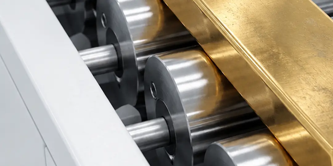 Close-up of a gold sheet being smoothed and polished by a machine with shiny metal rollers.
