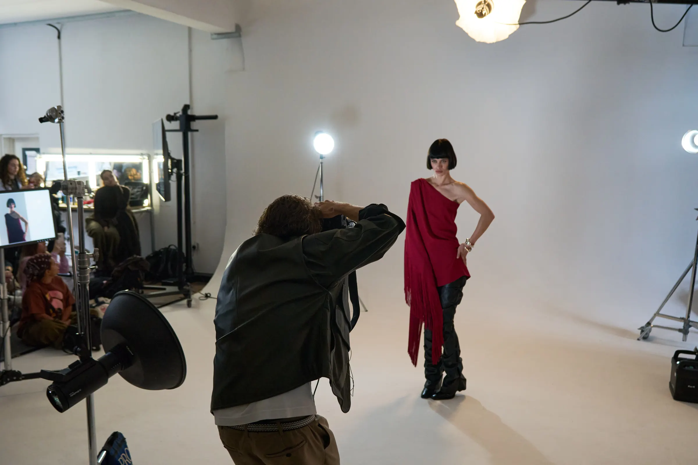A photographer shoots a model in a red one-shoulder dress on a white studio set while participants watch, at a Behind the Shoot masterclass