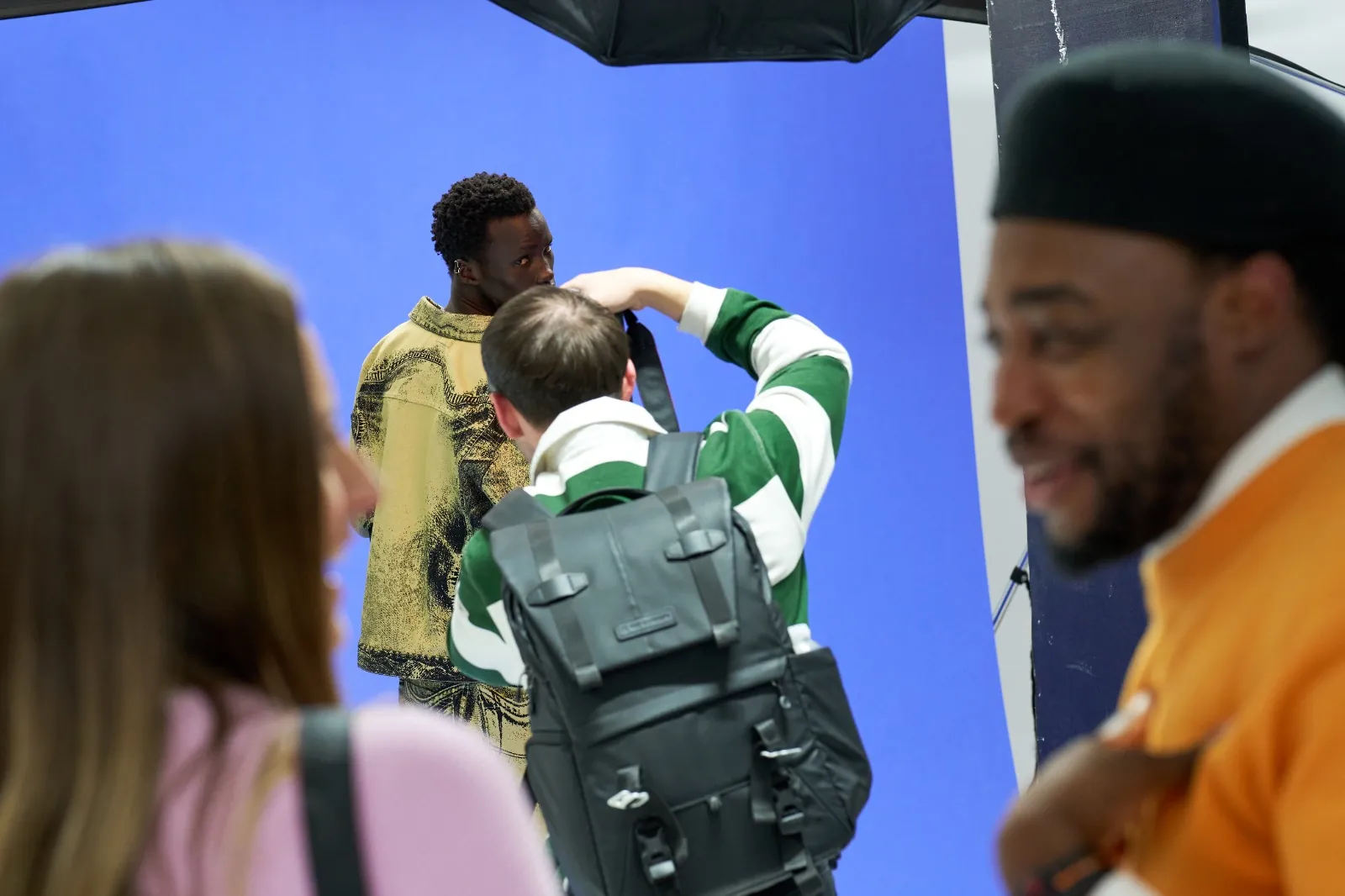 A participant photographs a model in a printed denim jacket against a blue backdrop while others talk on set at a Behind the Shoot event