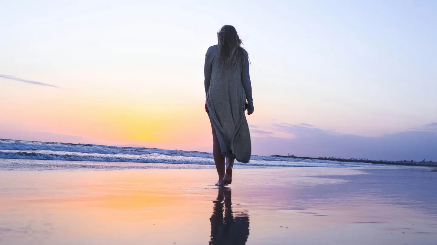 A woman walking alone along a beach at sunset, seen from behind