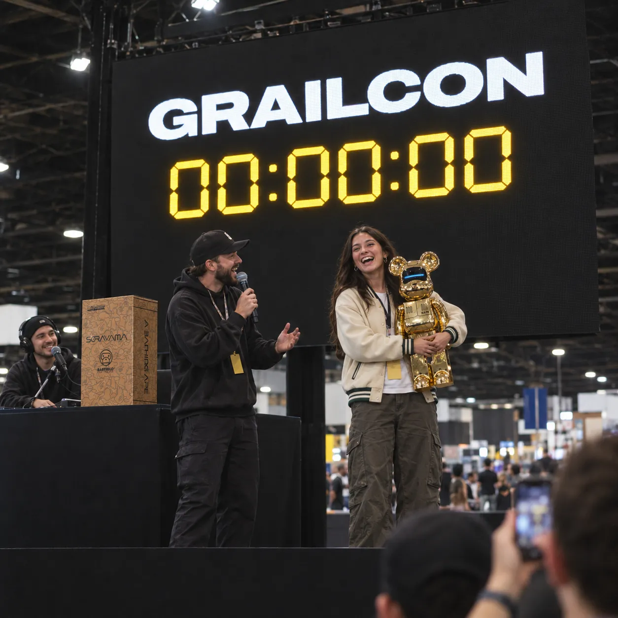 A giveaway winner on stage at a convention hugging a gold collectible figure, beside a host, under a screen showing a countdown at zero