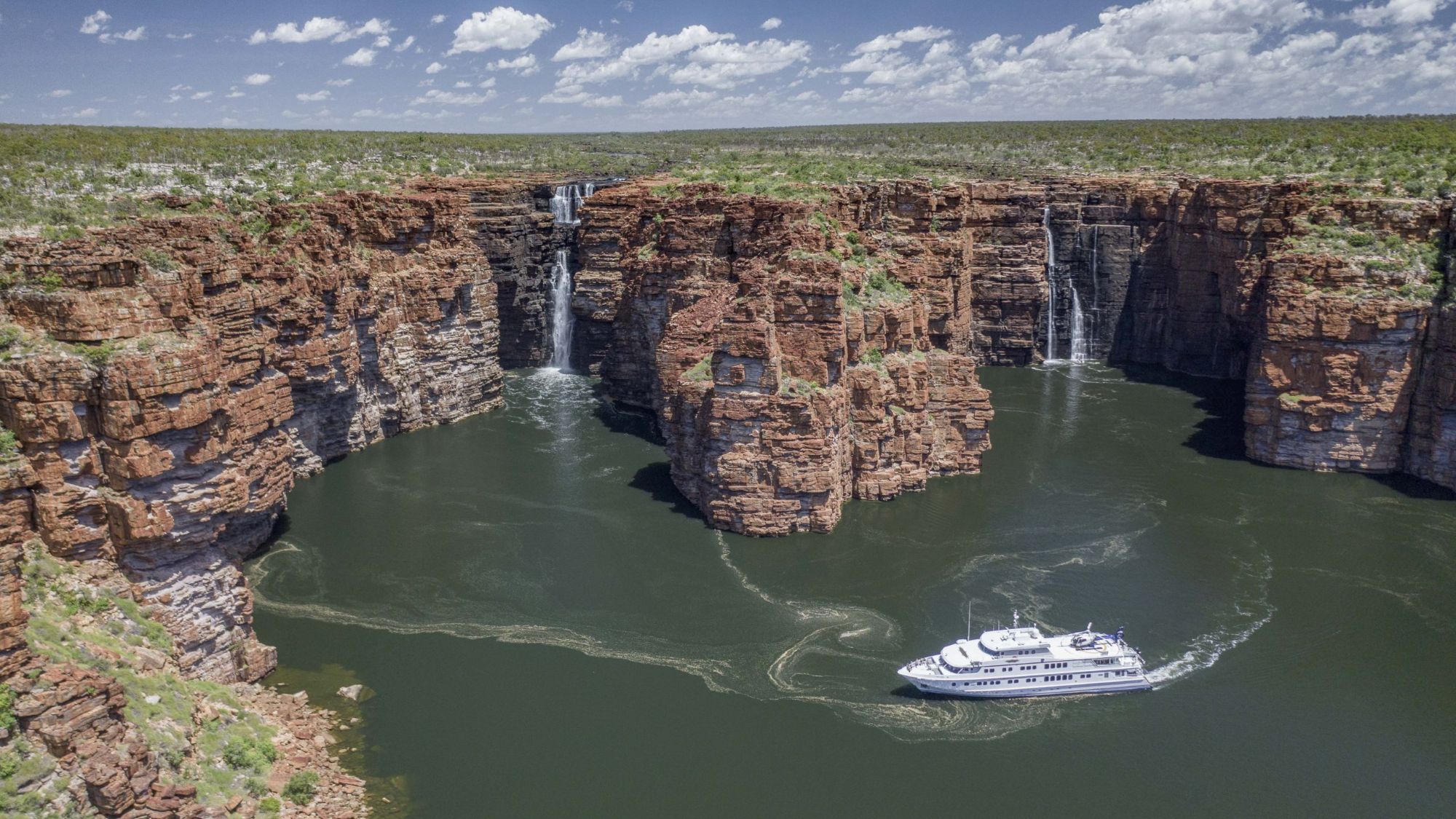 Expedition ship cruising beneath twin waterfalls in a red sandstone gorge in the Kimberley