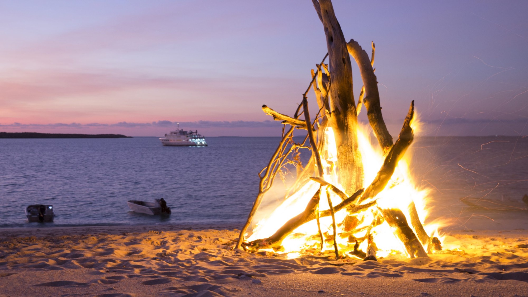 Beach bonfire at dusk with the ship anchored offshore