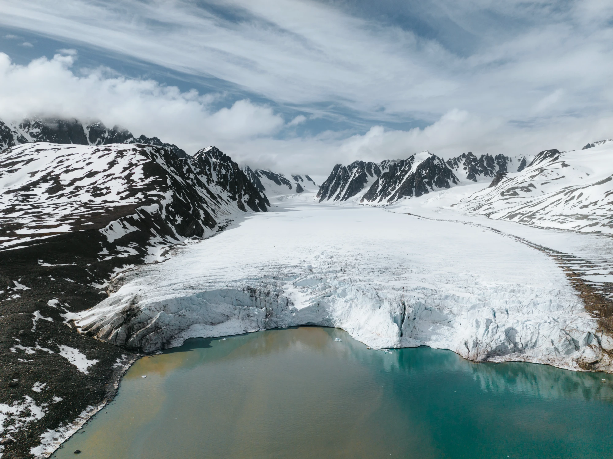 Aerial view of a glacier flowing between snow-capped peaks into a turquoise Svalbard fjord