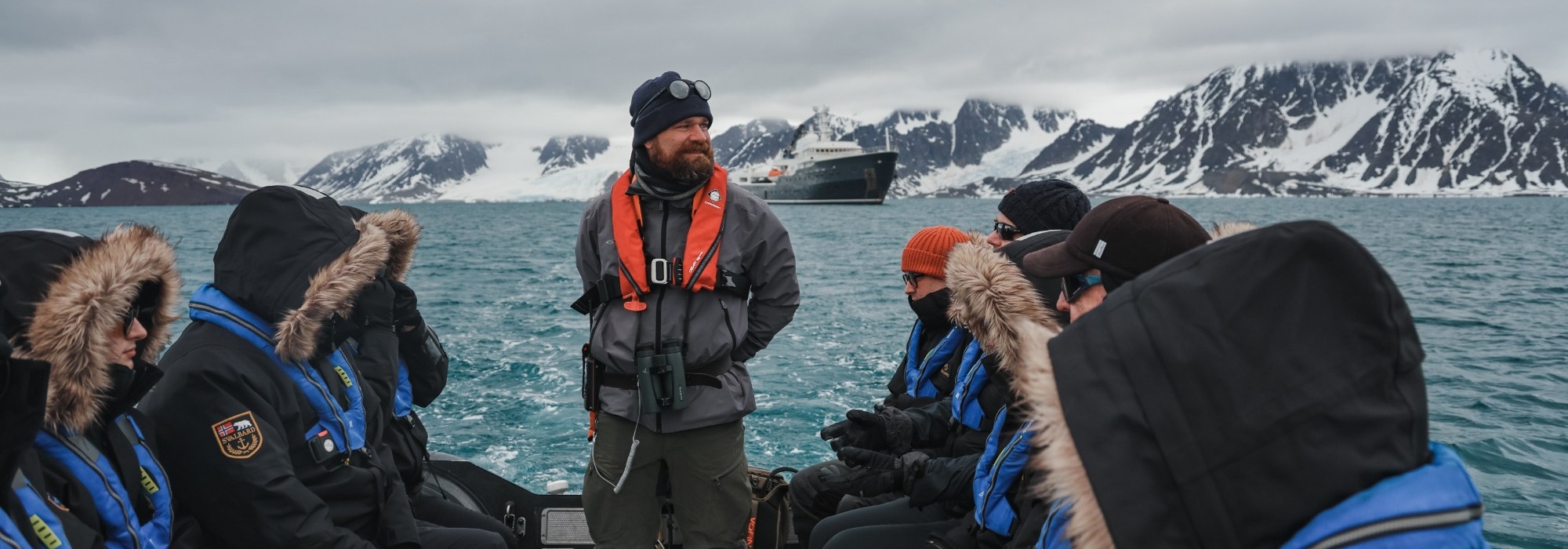 Expedition guide with binoculars in a Zodiac, with the ship and glaciers beyond