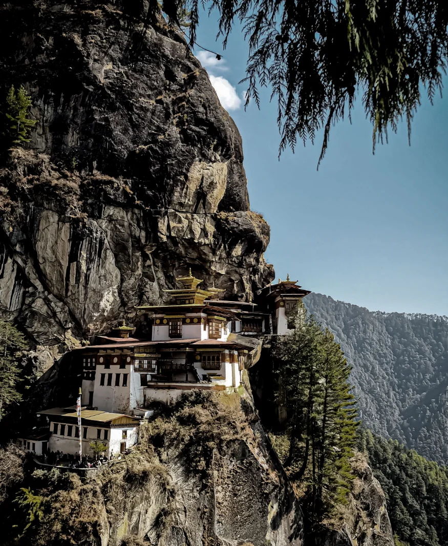 A white monastery built into the side of a steep cliff above a forested valley