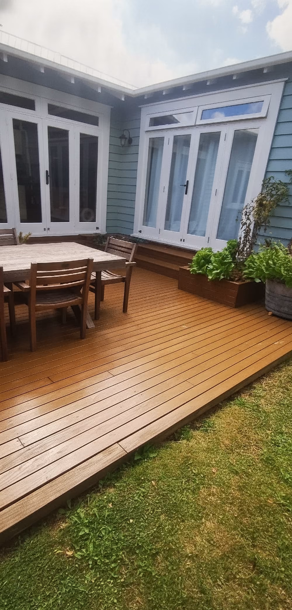 Wooden deck with a dining table and four chairs, adjacent to a house with light blue weatherboard siding and white-framed glass doors and windows, featuring potted plants and green lawn surrounding the deck.