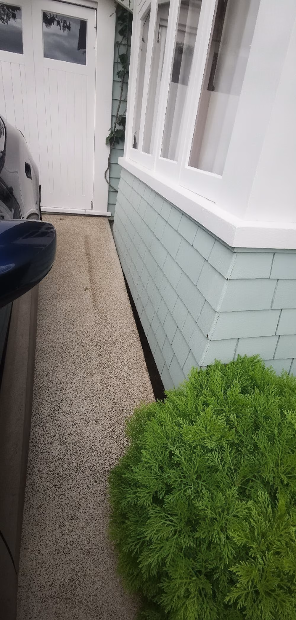 Narrow outdoor side walkway next to a house with pale green shingle siding and white-trimmed windows, a white door at the end, a parked car on the left, and a green bush in the foreground on the right.