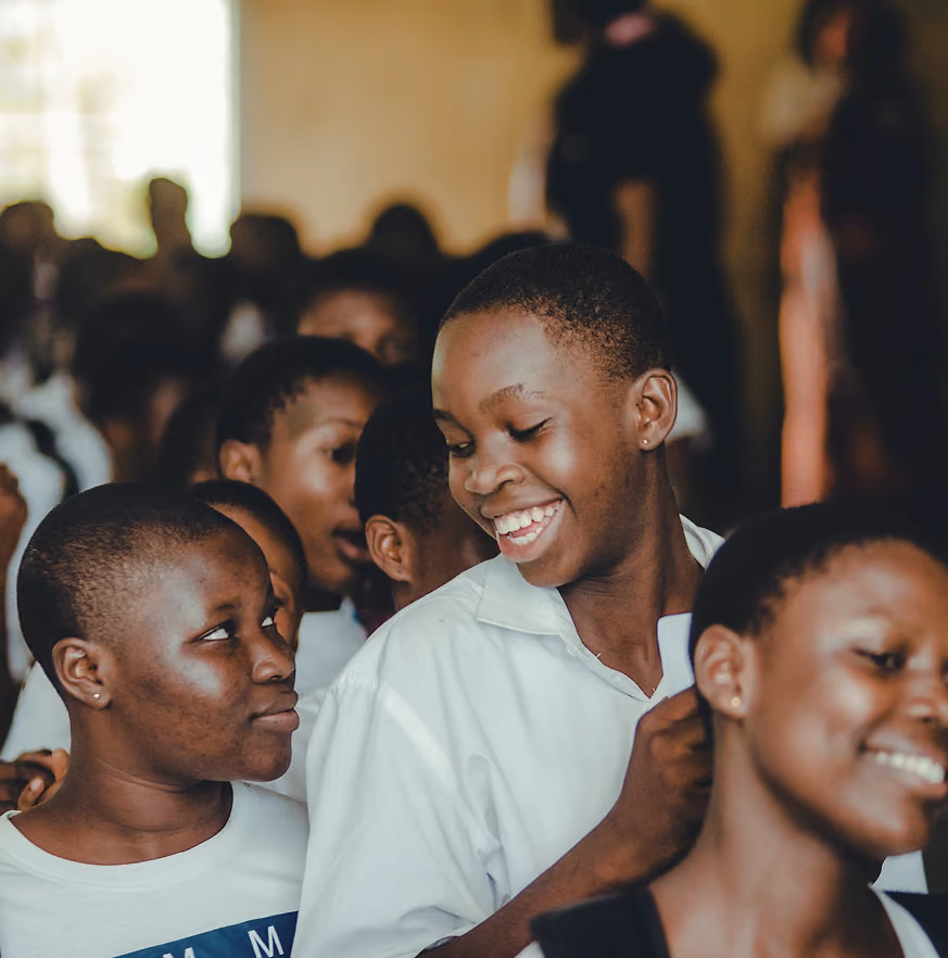 A group of smiling and happy young students in a crowded room, with two girls in the foreground engaging cheerfully with each other.