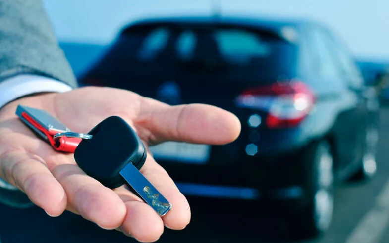 A hand holding out car keys in front of a parked car