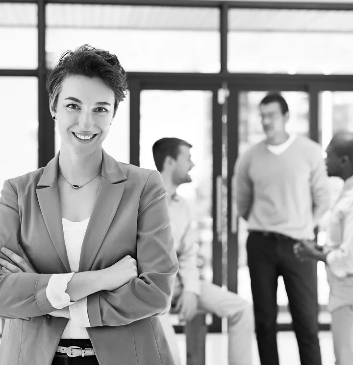 Confident young woman in business attire smiling with arms crossed in the foreground, with three colleagues casually talking in the background of a modern office setting.