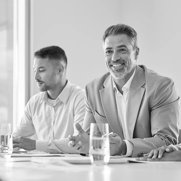 Smiling middle-aged man in a suit jacket speaking at a meeting table, with a younger man beside him looking downward, both seated with glasses of water on the table.