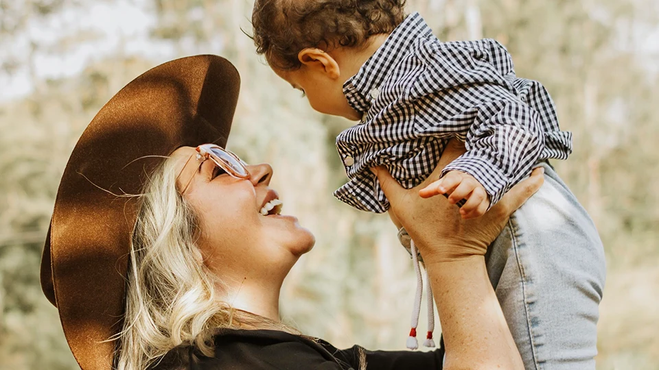 A woman in a wide brimmed hat laughing as she lifts a small child into the air outdoors.