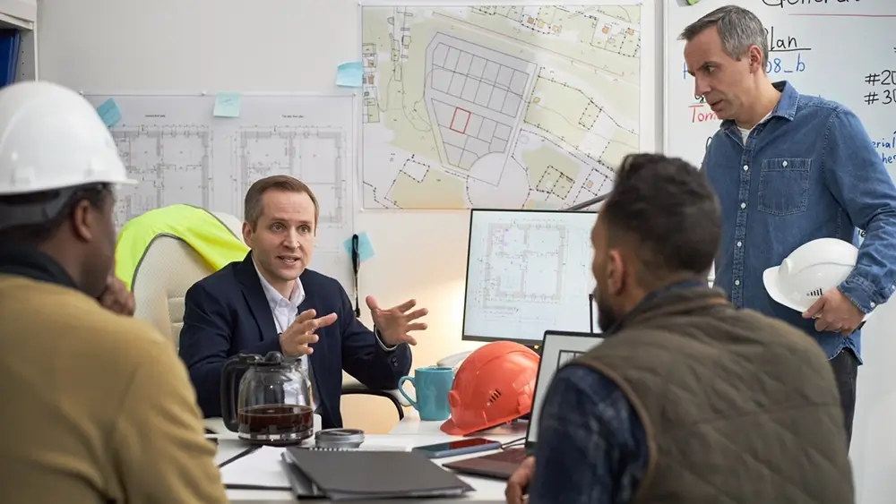 A manager talking with his team around a desk, hard hats and building plans beside them.