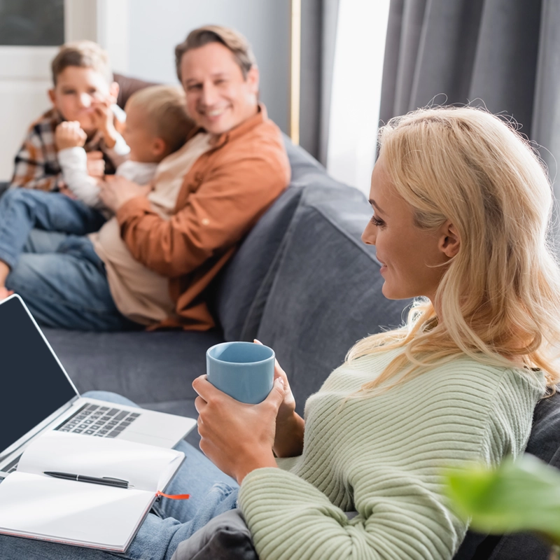 A mother with a coffee mug and an open laptop and notebook on the couch, smiling at her husband and two young sons beside her.