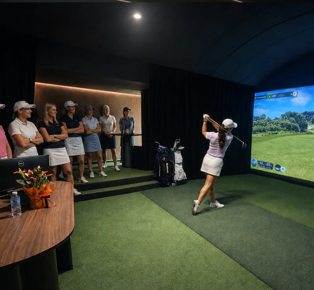 A woman swinging a golf club indoors while a group of women wearing golf attire and caps watch her in a room with green carpet and a large screen displaying a golf course simulation.