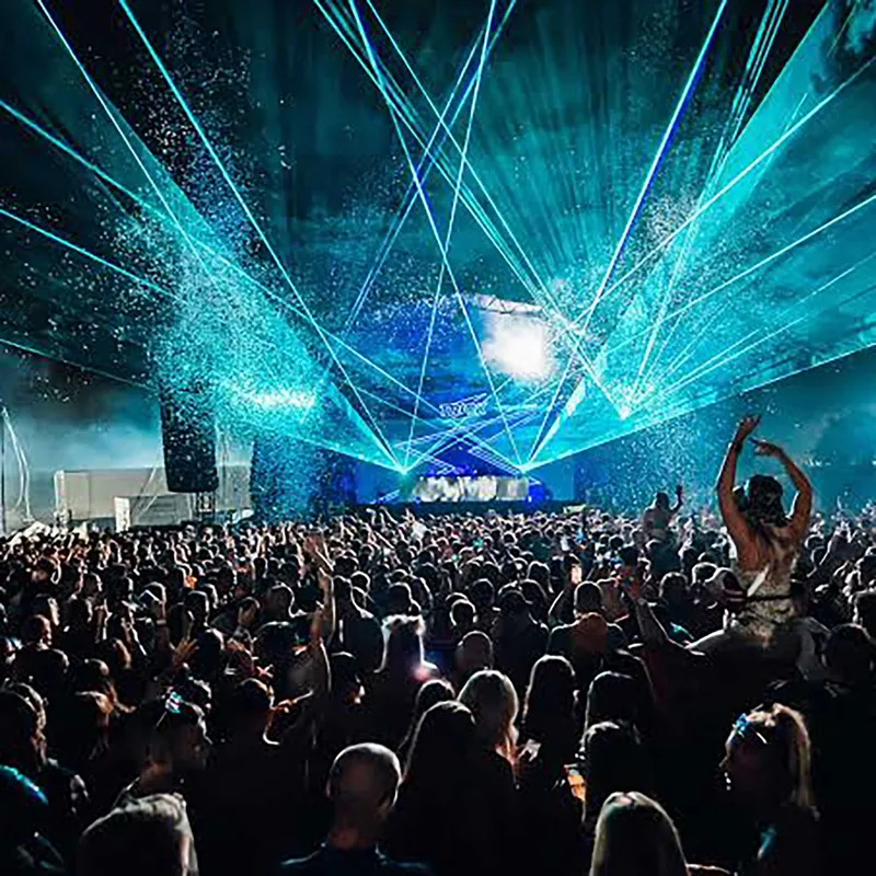 Large crowd at a nighttime music festival with blue laser lights and confetti illuminating the stage and audience.