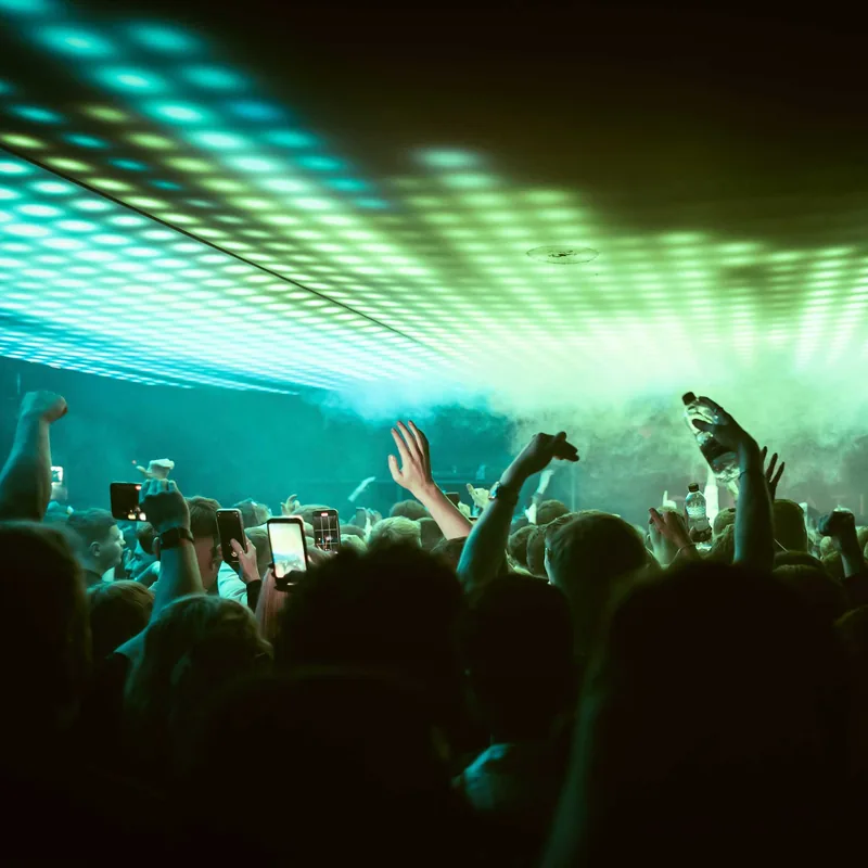 Crowd at a concert or event with raised hands and some people holding up smartphones, illuminated by green and blue stage lights on the ceiling.