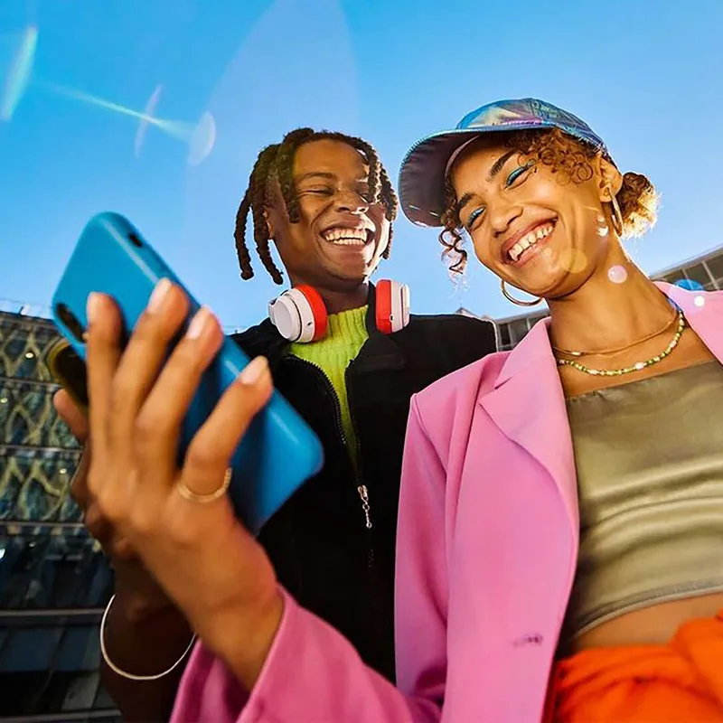 Two young adults outdoors smiling and looking at a blue smartphone, with a clear blue sky background; one wears red and white headphones around their neck and the other wears a shiny blue cap and pink jacket.