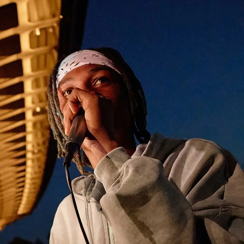 Young man with dreadlocks and a white bandana holding a microphone close to his mouth, wearing a gray hoodie, performing or speaking against a dark blue evening sky with a wooden ceiling structure on the left side.