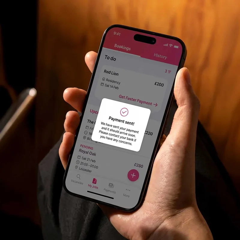 Hand holding a smartphone displaying a job booking app screen with a confirmation pop-up that says "Payment sent! We have sent your payment and it should arrive soon. Please contact your bank if you have any concerns." The screen lists jobs with details like location, date, time, and pay amounts.