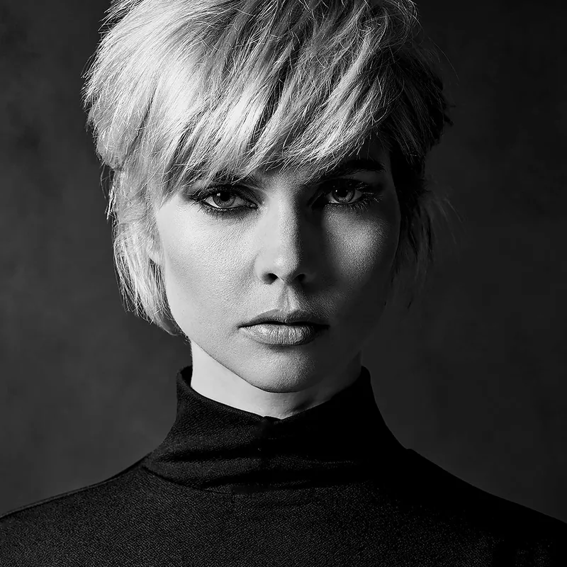 Black and white close-up portrait of a person with short, light-colored hair wearing a high-neck dark top, looking directly at the camera with a neutral expression.