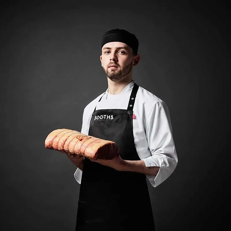 Man wearing a black chef hat, white shirt, and black apron with 'BOOTHS' holding a large, sliced roasted meat against a dark background.