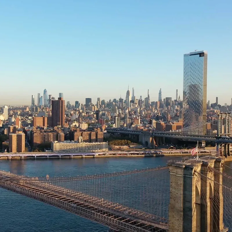 View of New York City skyline with the Brooklyn Bridge in the foreground and the Manhattan Bridge visible behind it, under a clear blue sky.