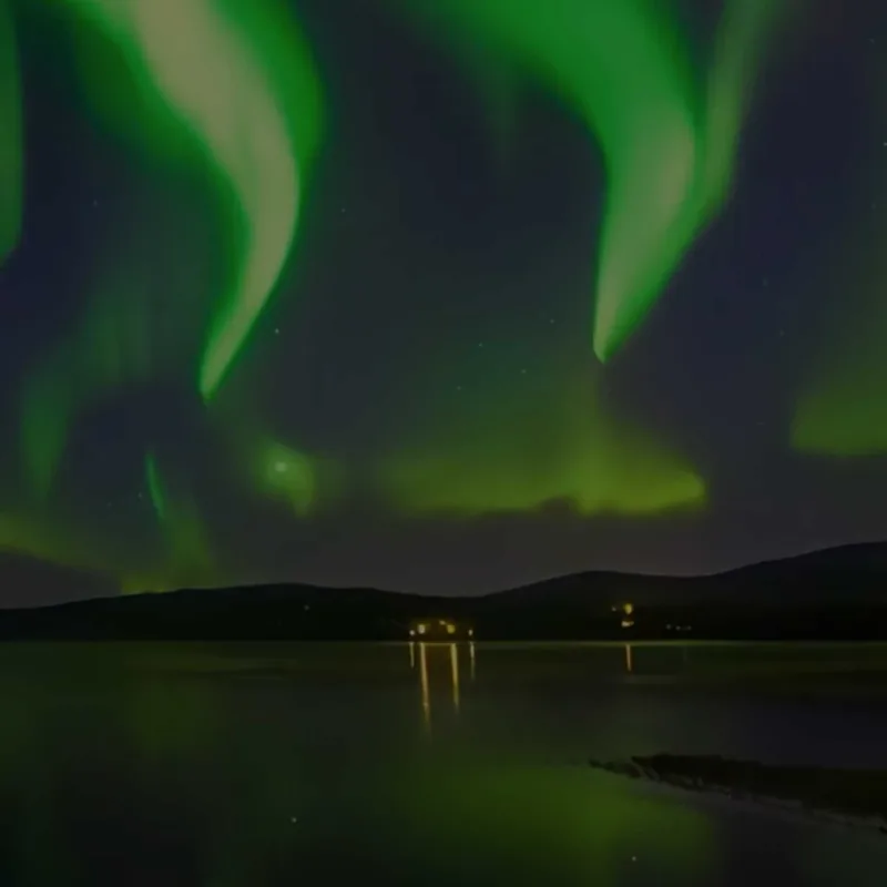 Green northern lights (aurora borealis) shining in waves across the night sky over a dark lake with hills and small illuminated buildings reflecting on the water.