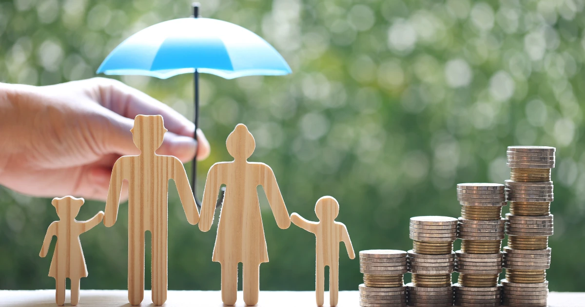 Wooden family figures protected by an umbrella beside stacks of coins