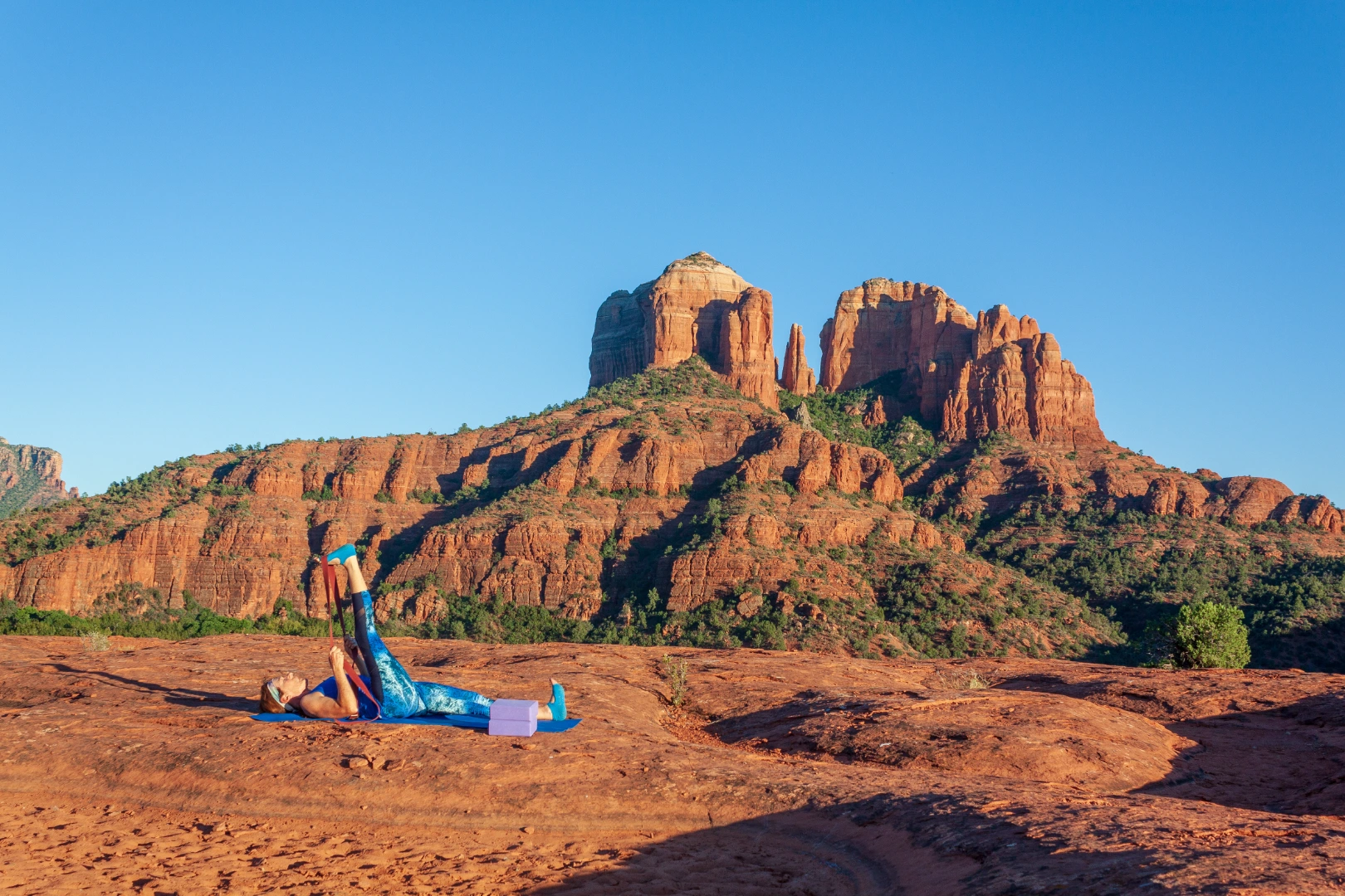 Person lying on a yoga mat doing leg raises on red rock with Cathedral Rock behind