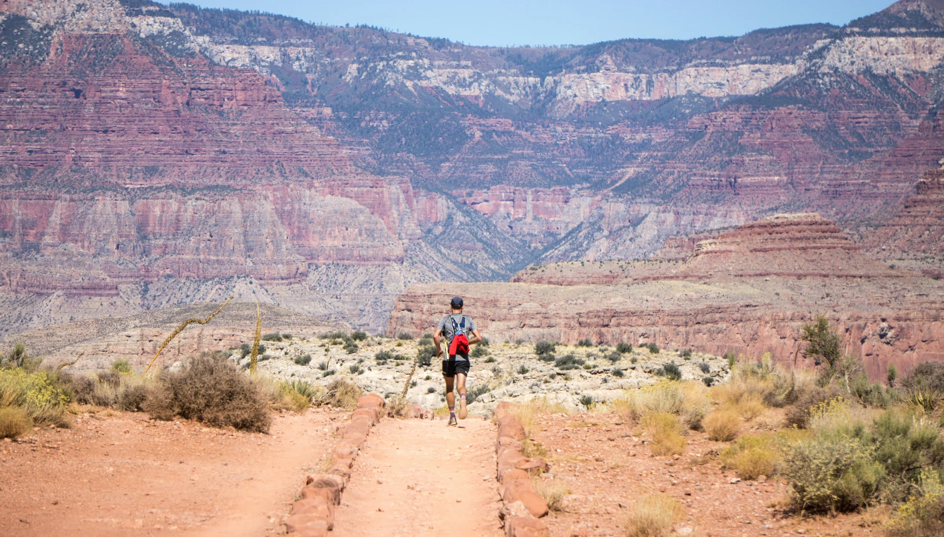 Hiker walking a dusty desert trail toward layered red canyon cliffs