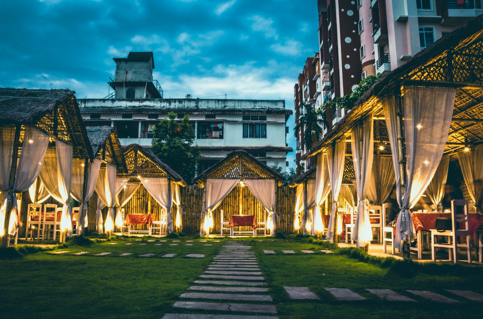 Evening courtyard with lit thatched pavilions and draped curtains, suggesting a relaxing place to stay