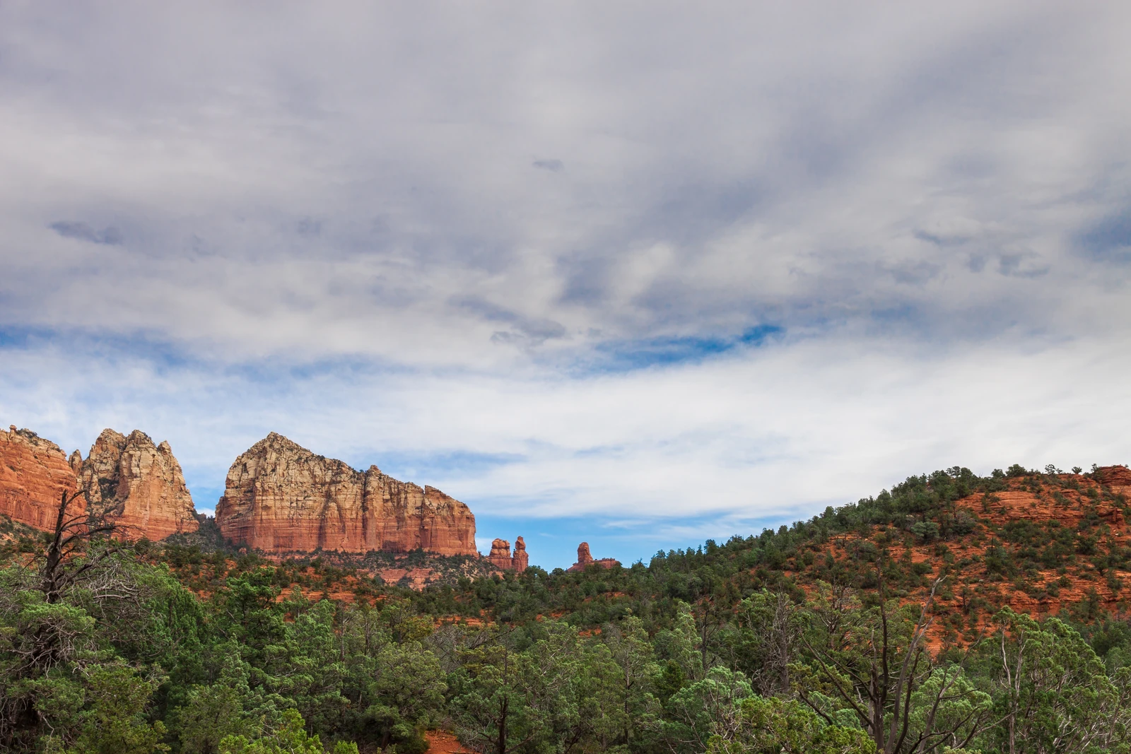 Red rock formations rising above green juniper forest under a broad cloudy sky