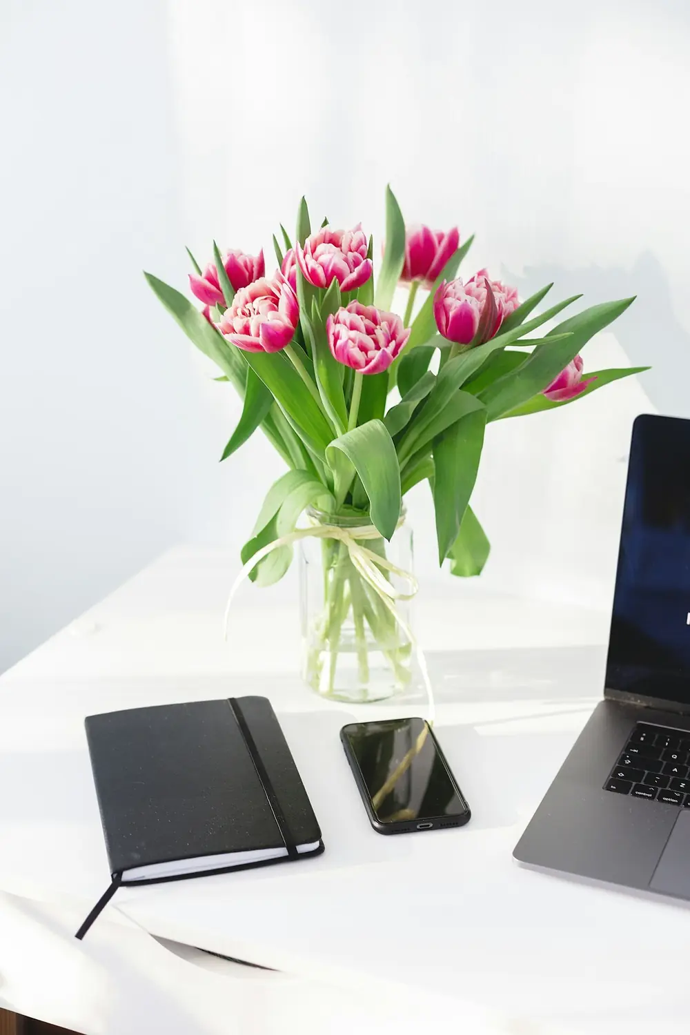 A vase of pink tulips on a white desk next to a laptop