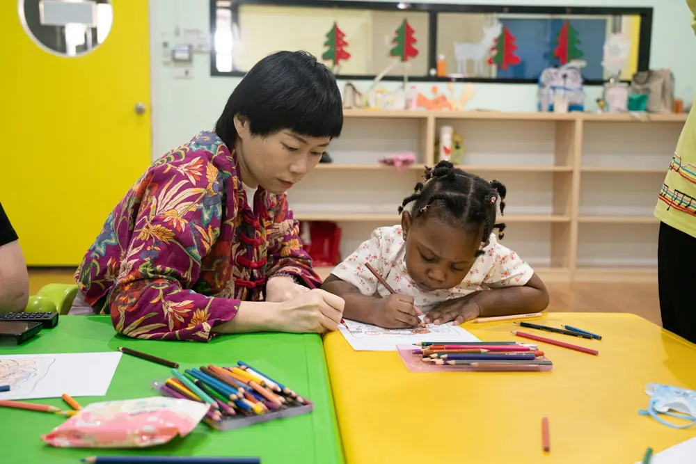 A teacher and a young student drawing together at a classroom table
