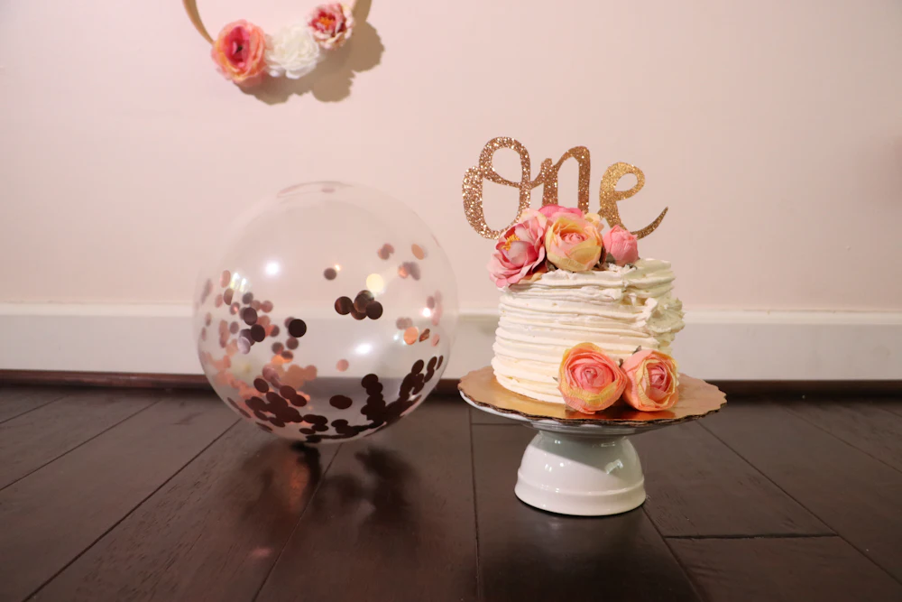 A small white first birthday cake topped with flowers and a gold One sign beside a confetti balloon