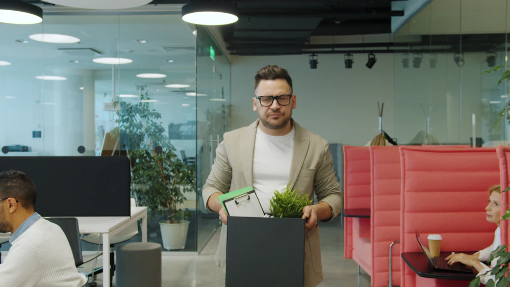 A smiling man carrying a box of his things and a small plant out of the office on his last day