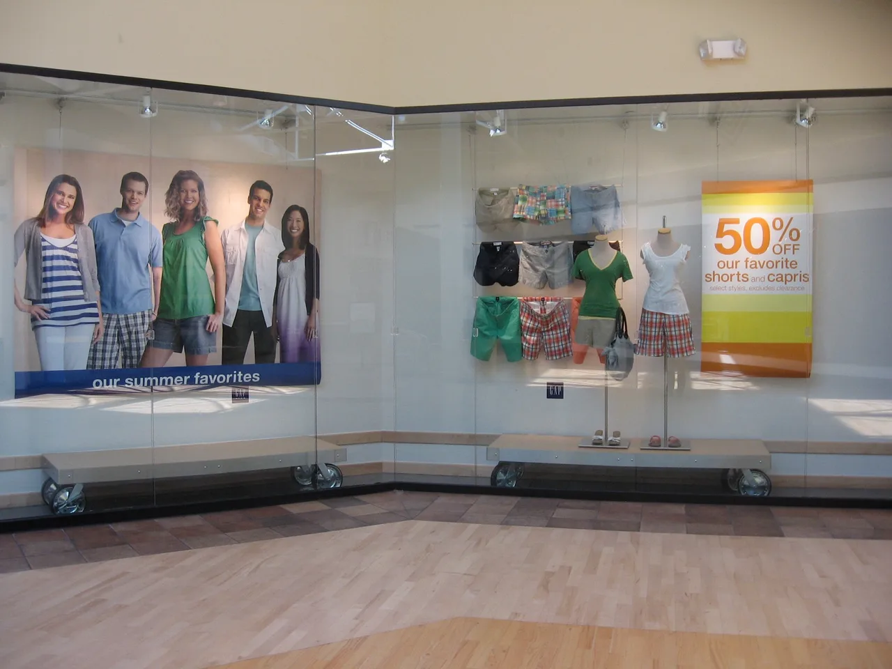 Store window display with five mannequins wearing summer shorts and tops, a 50% off sign for shorts and capris, and a banner showing five smiling people in summer clothes.