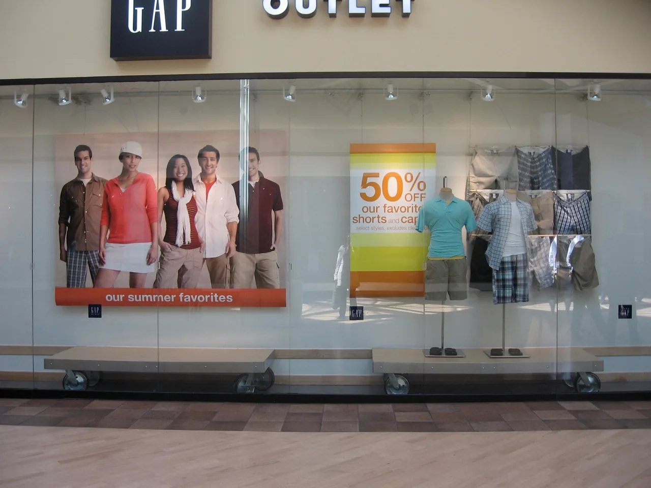 Storefront display at GAP Outlet featuring a poster of five young adults in summer clothing with a sign offering 50% off shorts and capris.