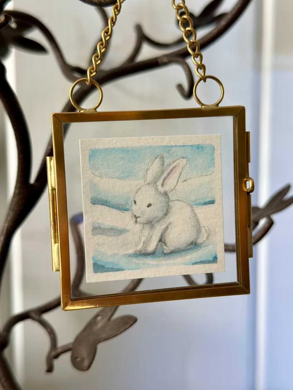 Small watercolor painting of a white snow bunny in a gold framed glass ornament hanging from a branch.