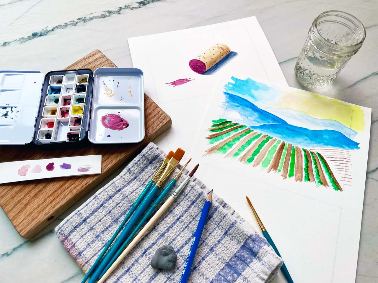 Watercolor painting setup with paint palette, brushes, pencil, cloth, glass of water, and paintings of a cork and a landscape with fields and hills.