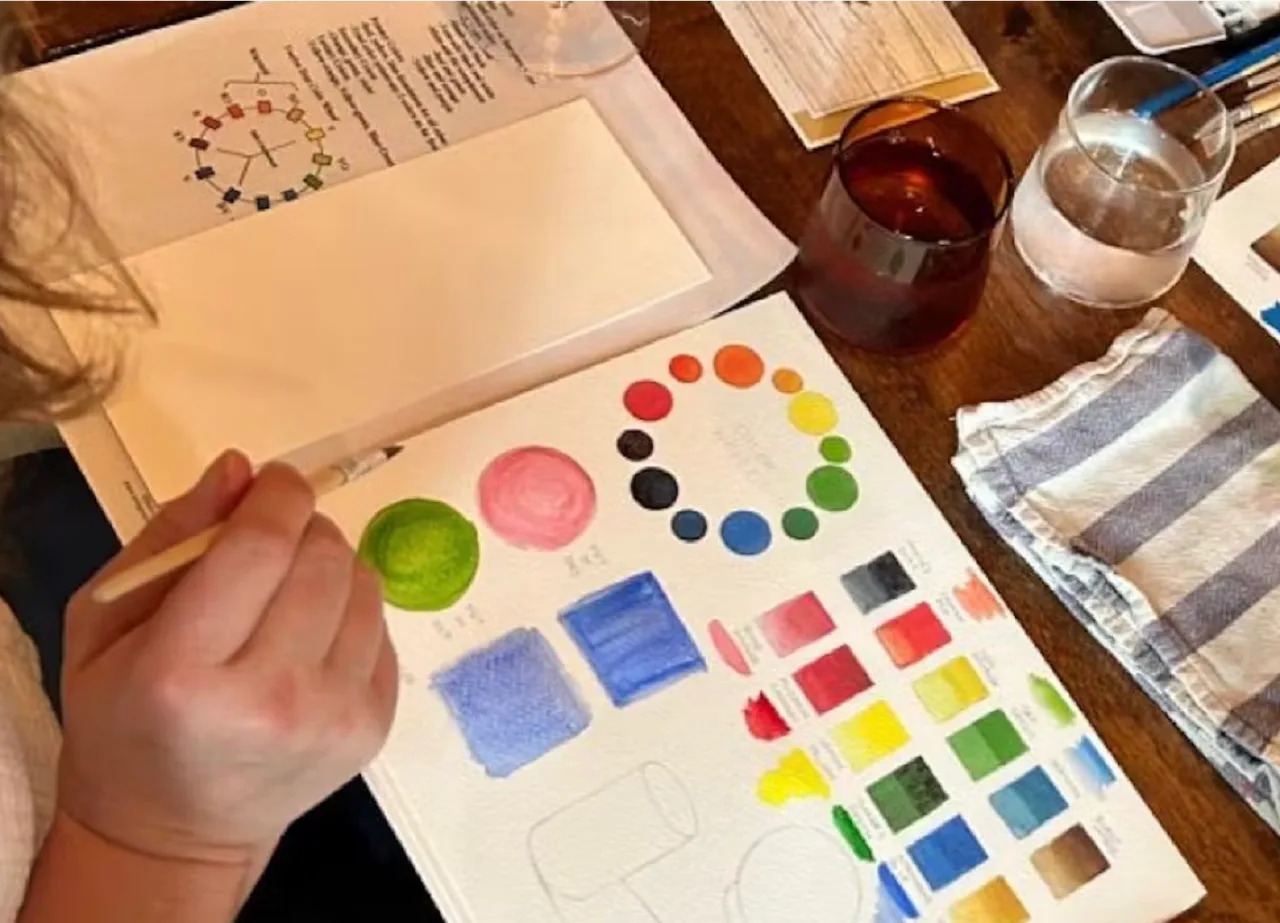 Person painting colorful circles, squares, and swatches on paper at a wooden table with glasses of water and a striped towel.