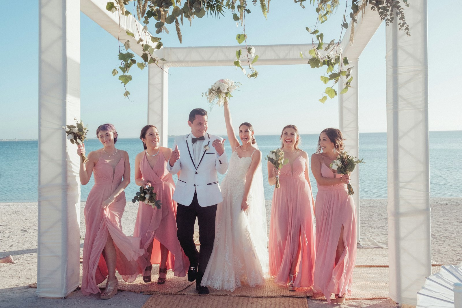 A bride, groom and bridesmaids celebrating under a beach wedding arch