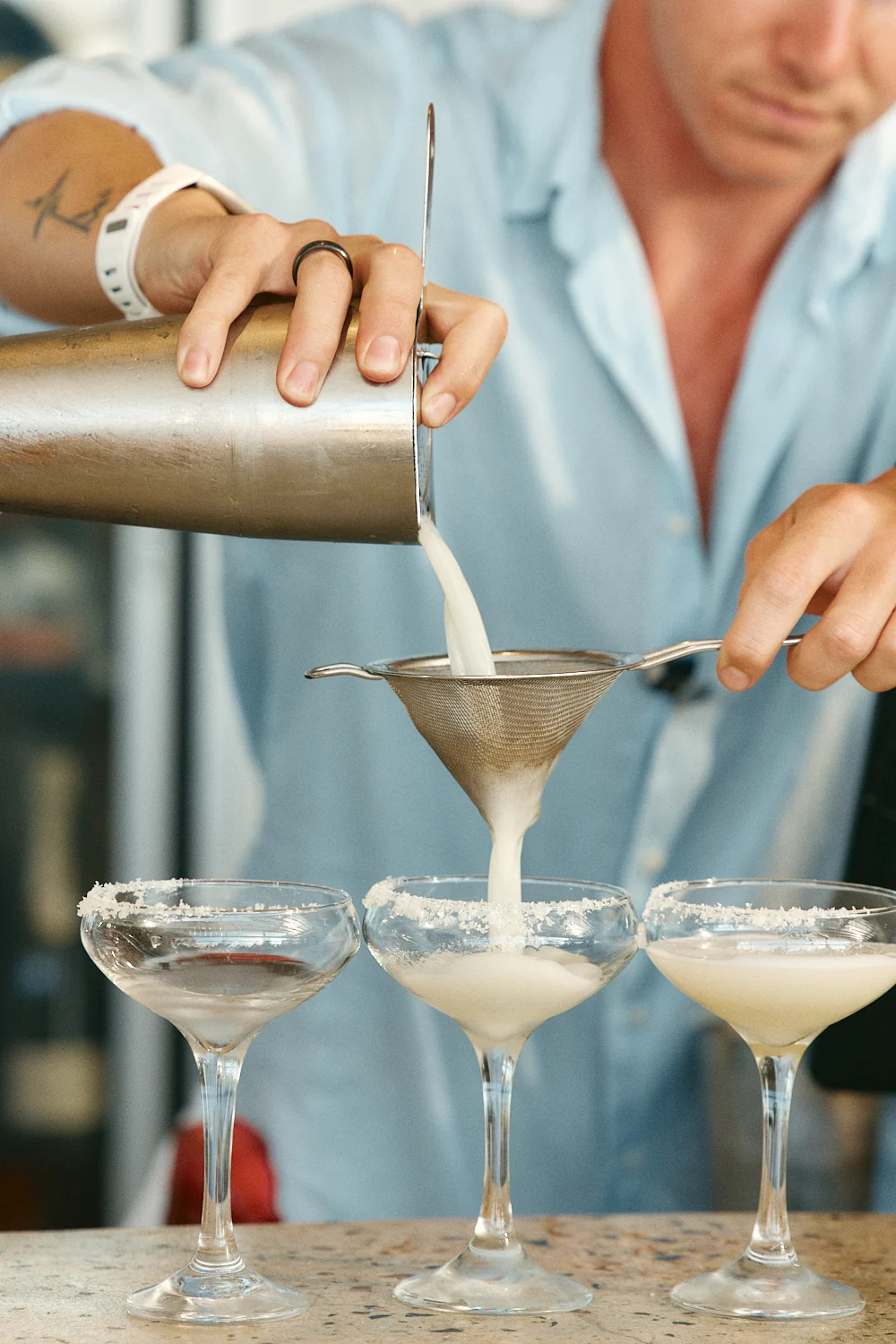 Bartender pouring cocktails into three salt-rimmed coupes