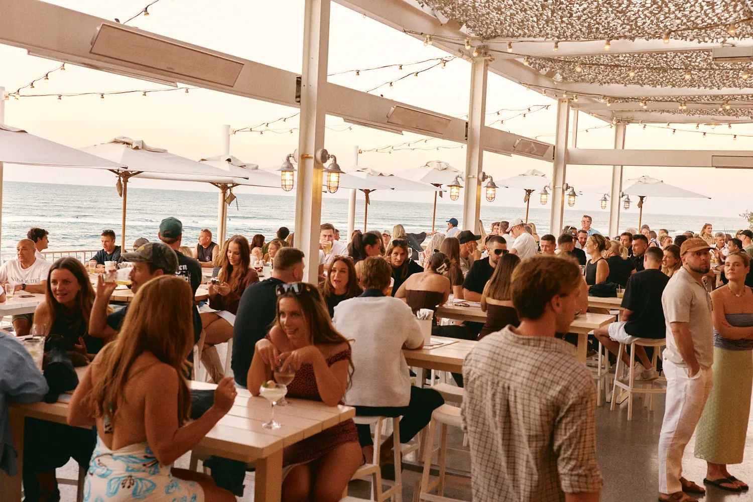 Busy oceanfront terrace at Burleigh Pavilion at sunset, with guests and festoon lights