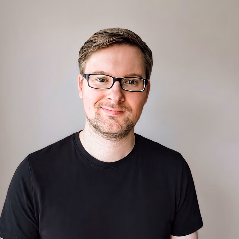 Porträt eines Mannes mit Brille und kurzem braunem Haar, der ein schwarzes T-Shirt trägt und leicht lächelt vor einem neutralen hellen Hintergrund.