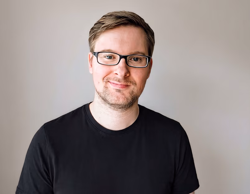 Bild von Tim Willer - Ein lächelnder Mann mit kurzen braunen Haaren, Brille und schwarzem T-Shirt vor hellem Hintergrund.