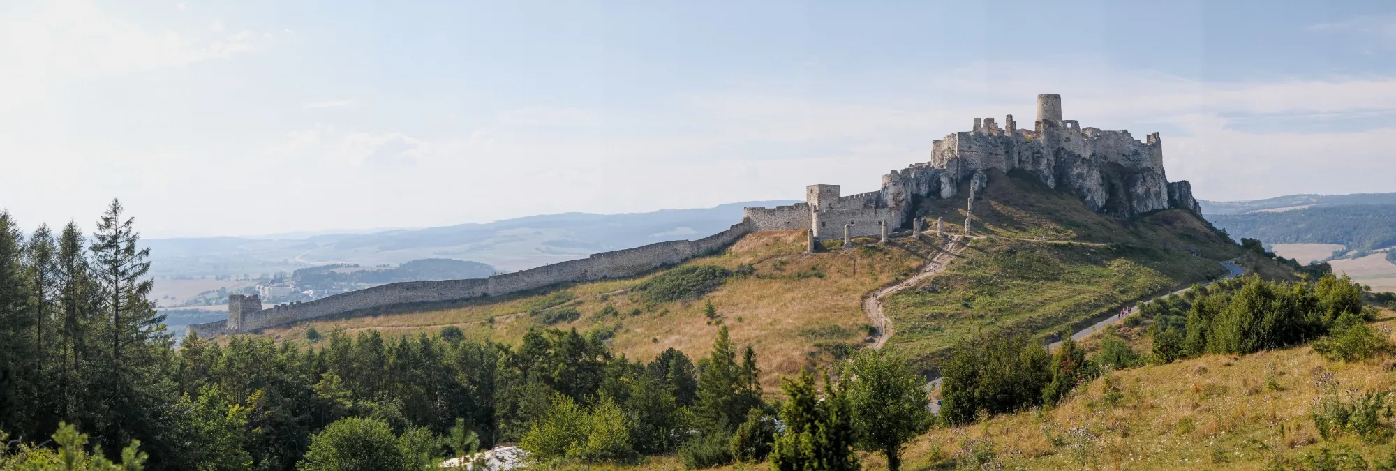 Panorama of Spiš Castle's ruined white walls and towers on a grassy hilltop, with a long fortified wall and forested valley below.