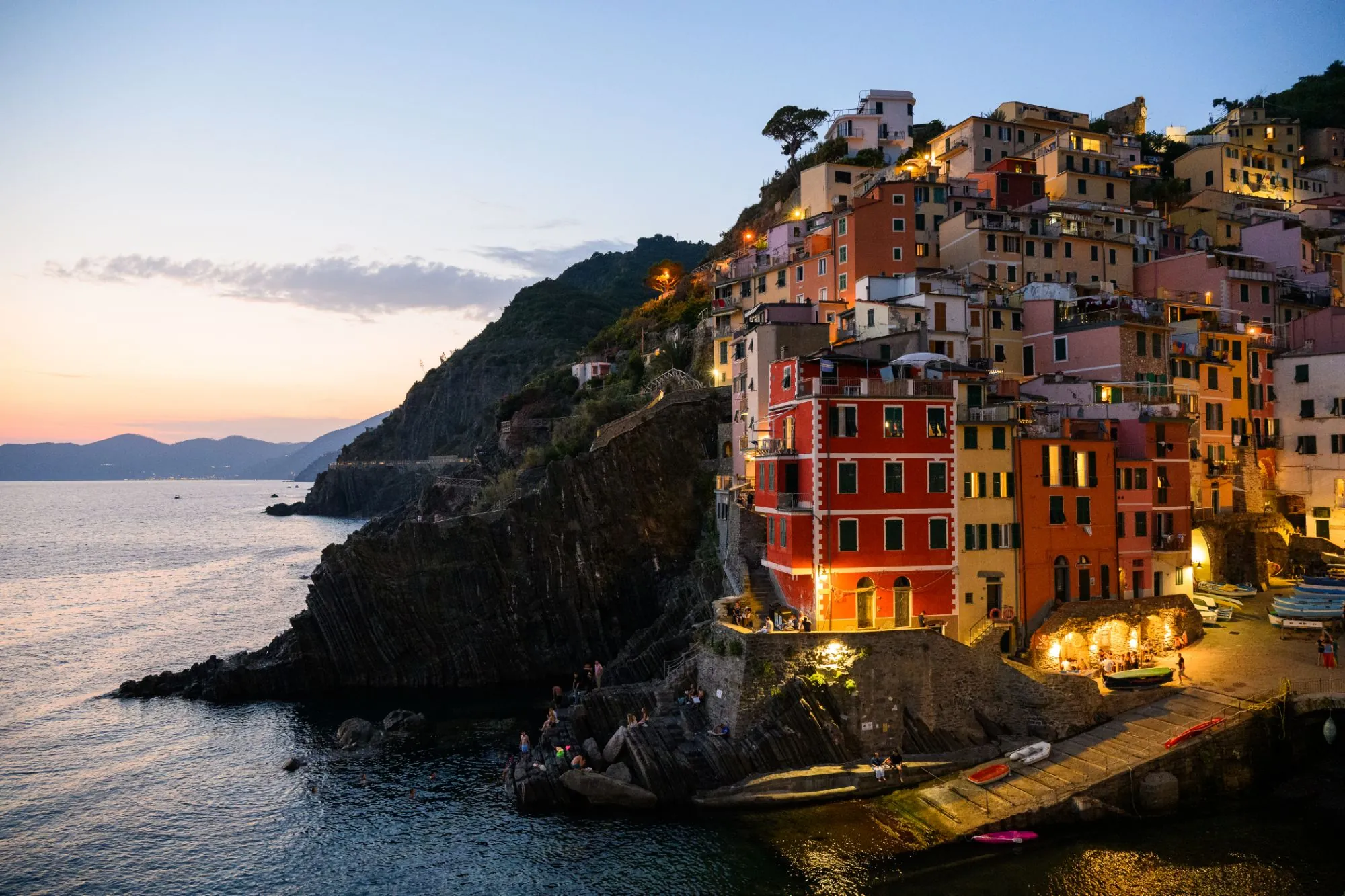 Colourful houses of Riomaggiore stacked on a cliff above the sea at dusk, with windows and harbour lights glowing.